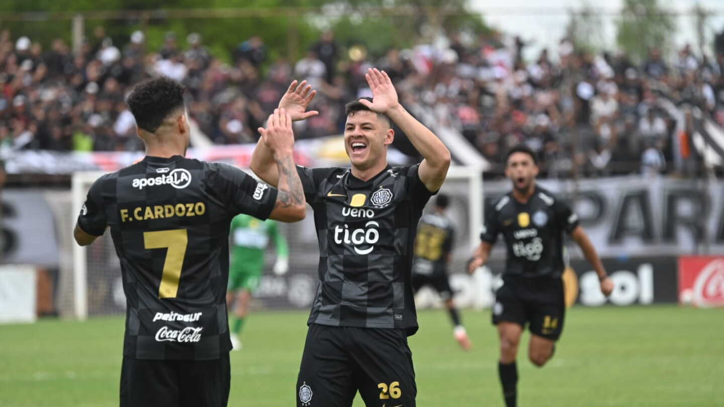 Dos jugadores de Olimpia, con camiseta negra a cuadros, chocan las manos para festejar un gol, con la hinchada y un arquero rival de verde al fondo.